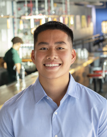 Headshot of a young Asian-American man looking at the camera smiling and wearing a blue button up shirt standing in front of a restaurant counter.